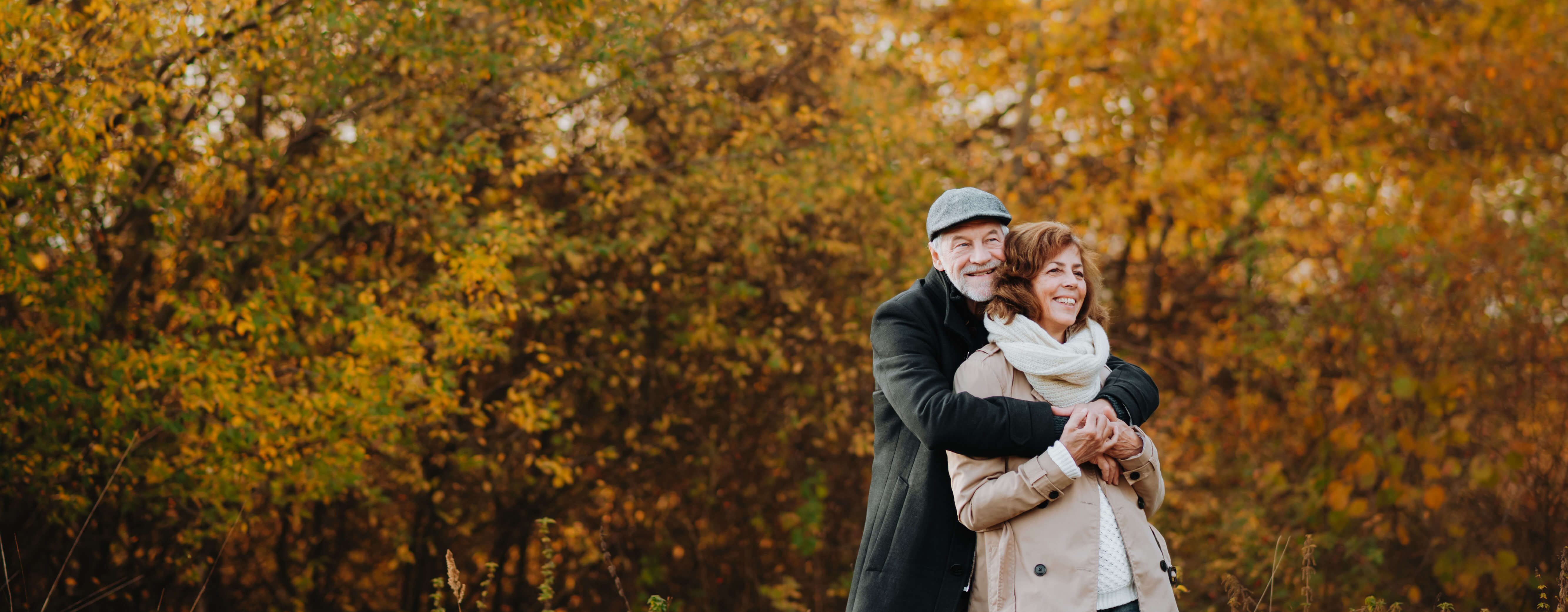 A couple of older people embracing while standing in a forest with orange autumnal trees.