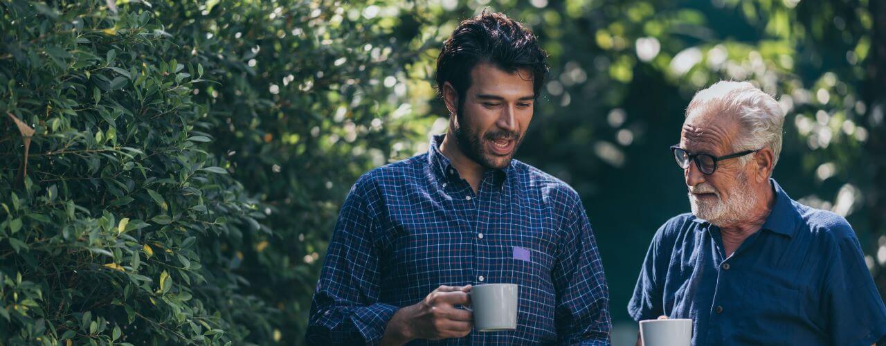 An older and younger person standing together in a forest and taking to one another while holding mugs of coffee.