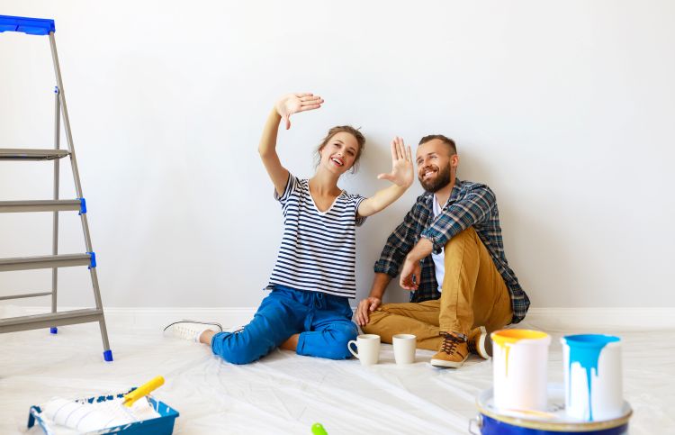A young couple sitting in an empty white room with cans of paint, a paint roller, and ladder around them. One is making a motion to frame a section of the wall with their hands and the other is smiling and looking toward where they're framing.
