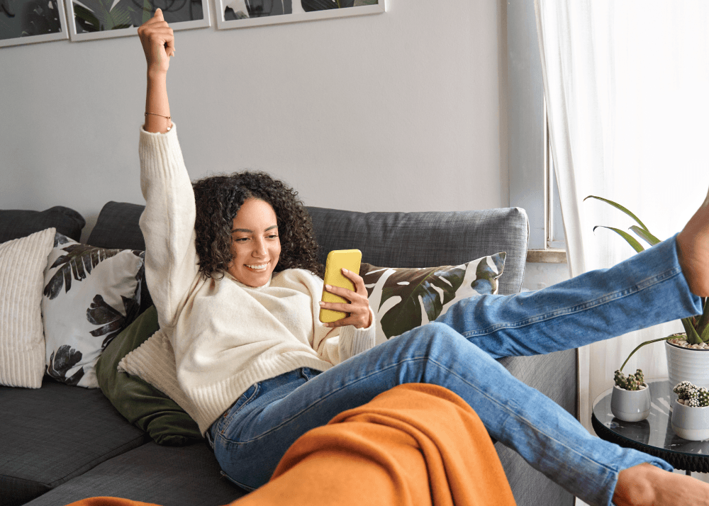 Woman leaning back onto her couch. She has one arm raised as she's cheering while looking at her phone in the other hand.