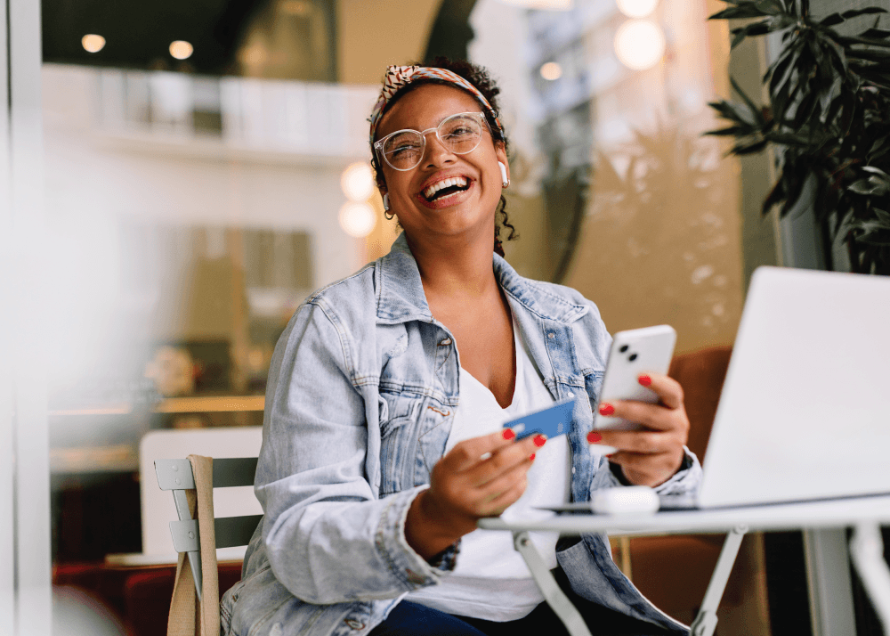 Woman sitting at a coffee shop with a laptop on the table in front of her. She is smiling while holding a debit card in one hand and her phone in the other.